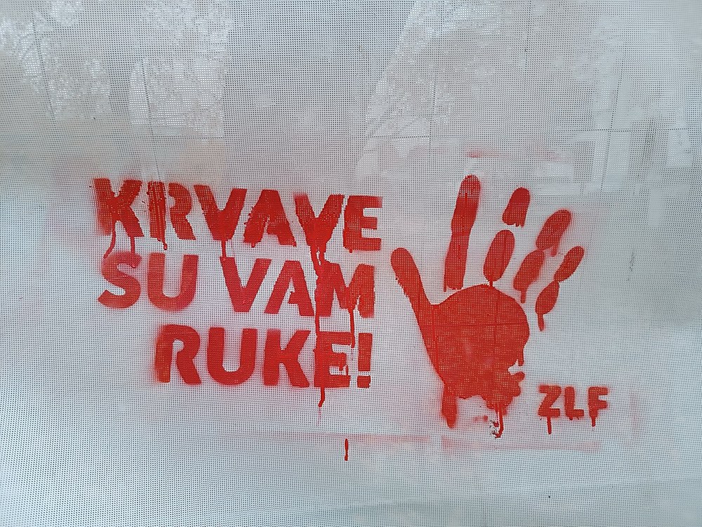 Црвени отисак шаке и натпис „Krvave su vam ruke!“ на белом платну.